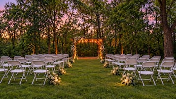 An outdoor wedding venue with rows of chairs arranged for guests, a floral archway, and a natural backdrop of trees.