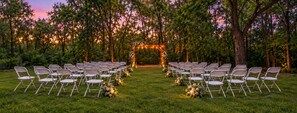 Un espacio para bodas al aire libre con filas de sillas dispuestas para los invitados, un arco floral y un entorno natural de árboles.