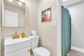 Et badeværelse med en hvid vask, guldfarvede armaturer, et hvidt toilet, et indrammet billede på væggen og en grøn bruseforhæng.