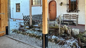Puinen rakennelma, jossa on katettu sisäänkäynti, kiviseinä, puuovi ja metallirunkoon kiinnitetty penkki.