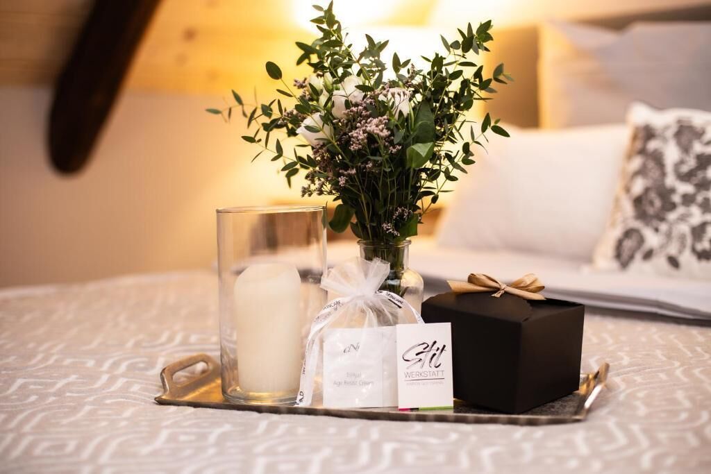 Ein Tablett mit Kerze, eine Glasvase mit Blumen, eine Geschenkbox und einer Karte auf einem Bett.