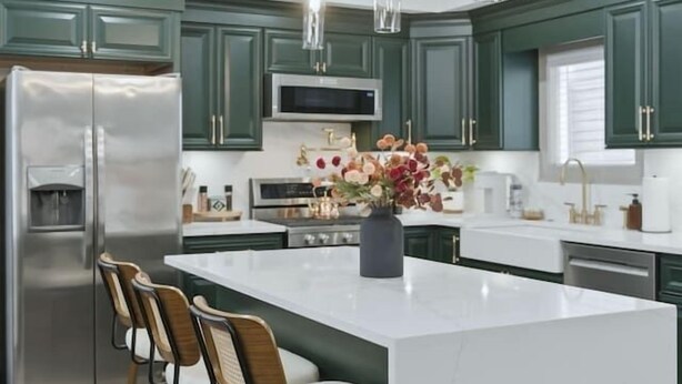 Cozinha moderna com armários verdes, eletrodomésticos de aço inoxidável, ilha branca com banquetas e iluminação pendente.