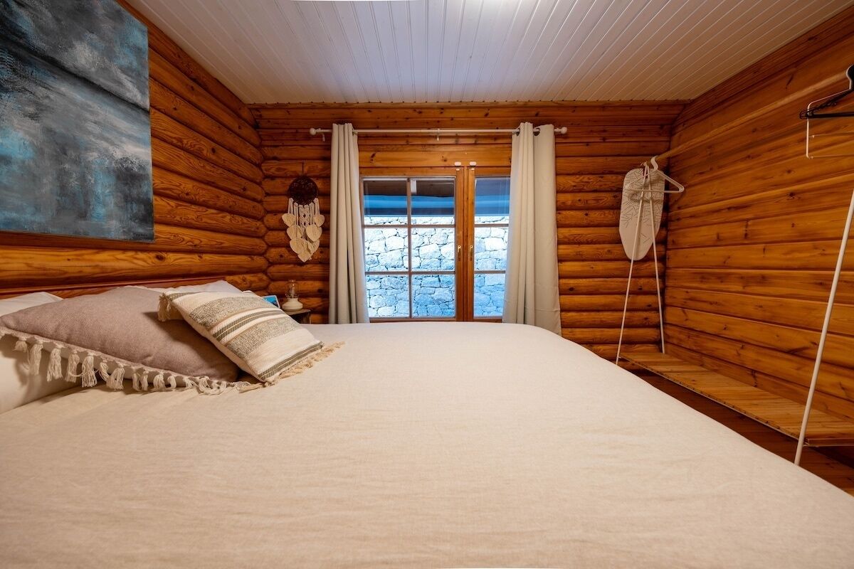 Un dormitorio tipo cabaña de troncos con cama, cortinas y una pared de piedra visible a través de la ventana.