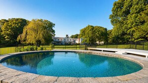 Une piscine aux eaux cristallines, bordée de pierres, entourée d’une clôture noire, avec un bâtiment visible au loin.