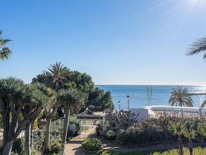 A coastal view with a pathway leading to a white building, surrounded by palm trees and various plants. - Apartment in Playas del Duque for 6 people | Casa Sevilla 511 (Puerto Banús - Marbella)