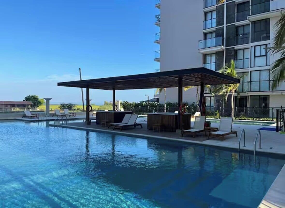 A swimming pool with lounge chairs and a covered seating area, surrounded by a modern building with balconies.