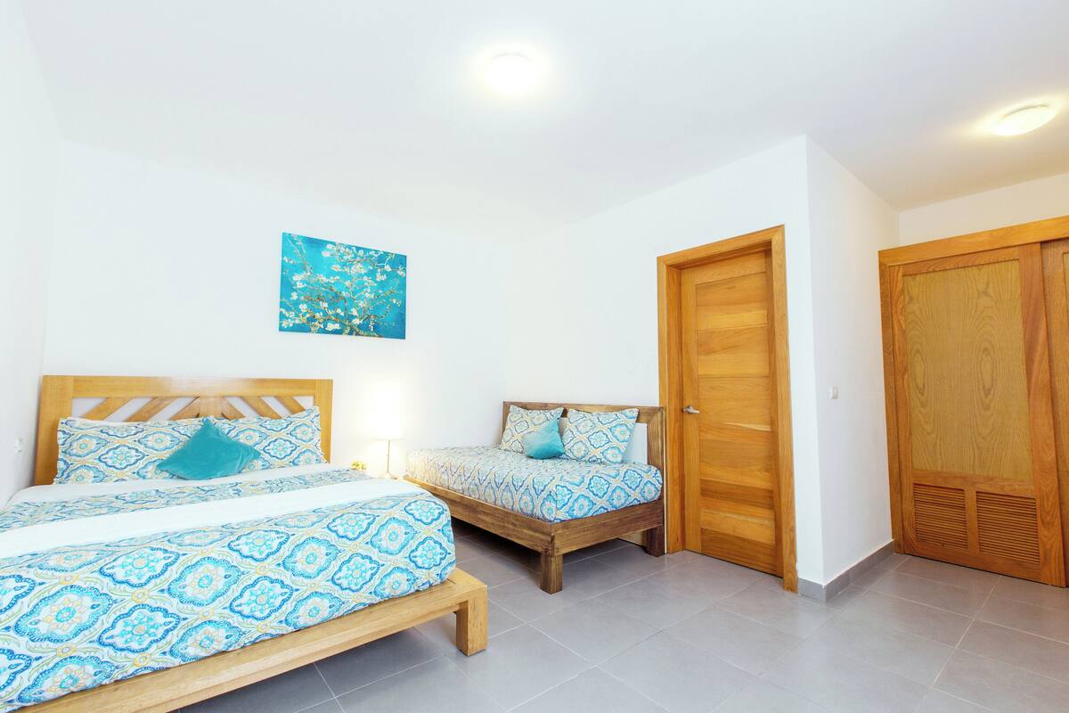 A bedroom with two beds, wooden headboards, a blue and white patterned bedspread, a small painting on the wall, and a wooden wardrobe.
