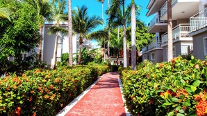 A pathway flanked by lush greenery and palm trees, leading to a building with balconies and a clear blue sky. - BRISAS TROPICAL playa LOS CORALES (Punta Cana)