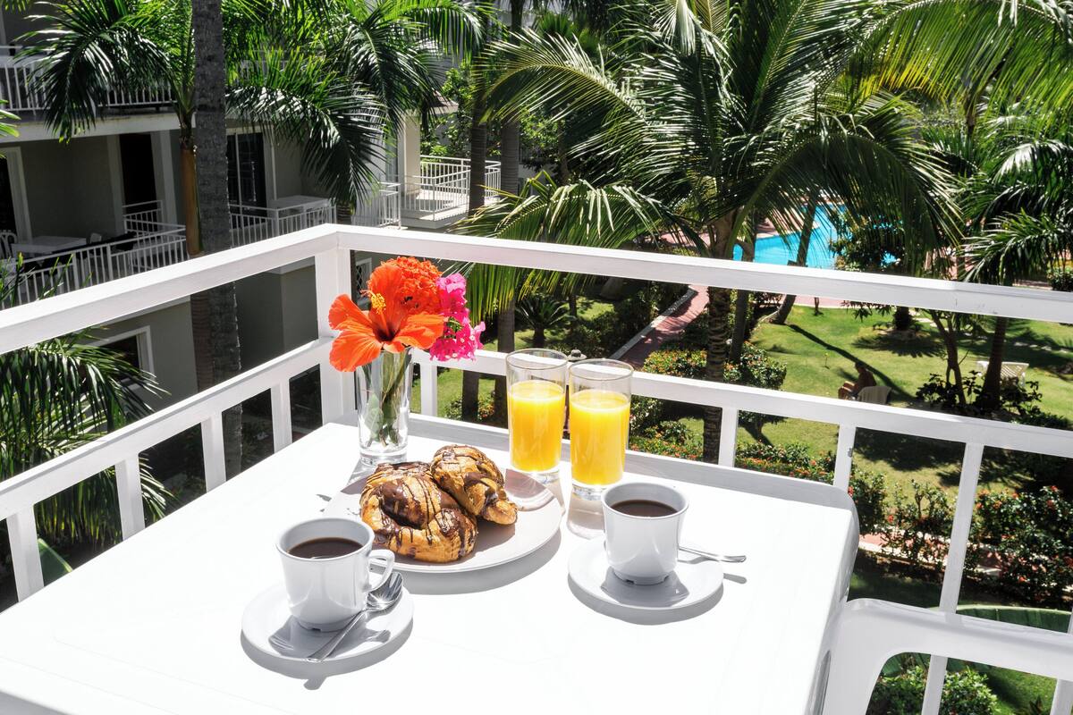 A balcony with a white table set for breakfast, including croissants, coffee, and orange juice, overlooking a pool and lush greenery.
