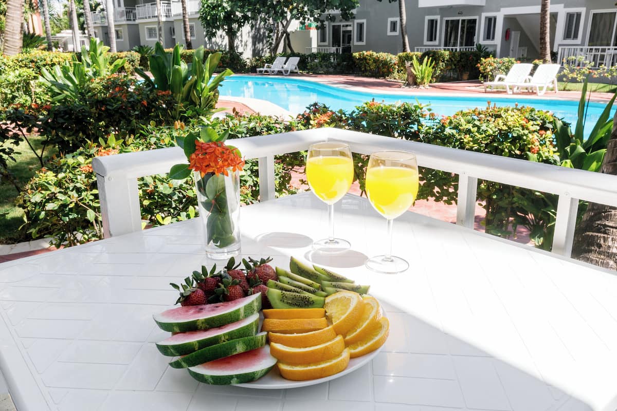 A table with fruit and drinks, a pool, and a building in the background.