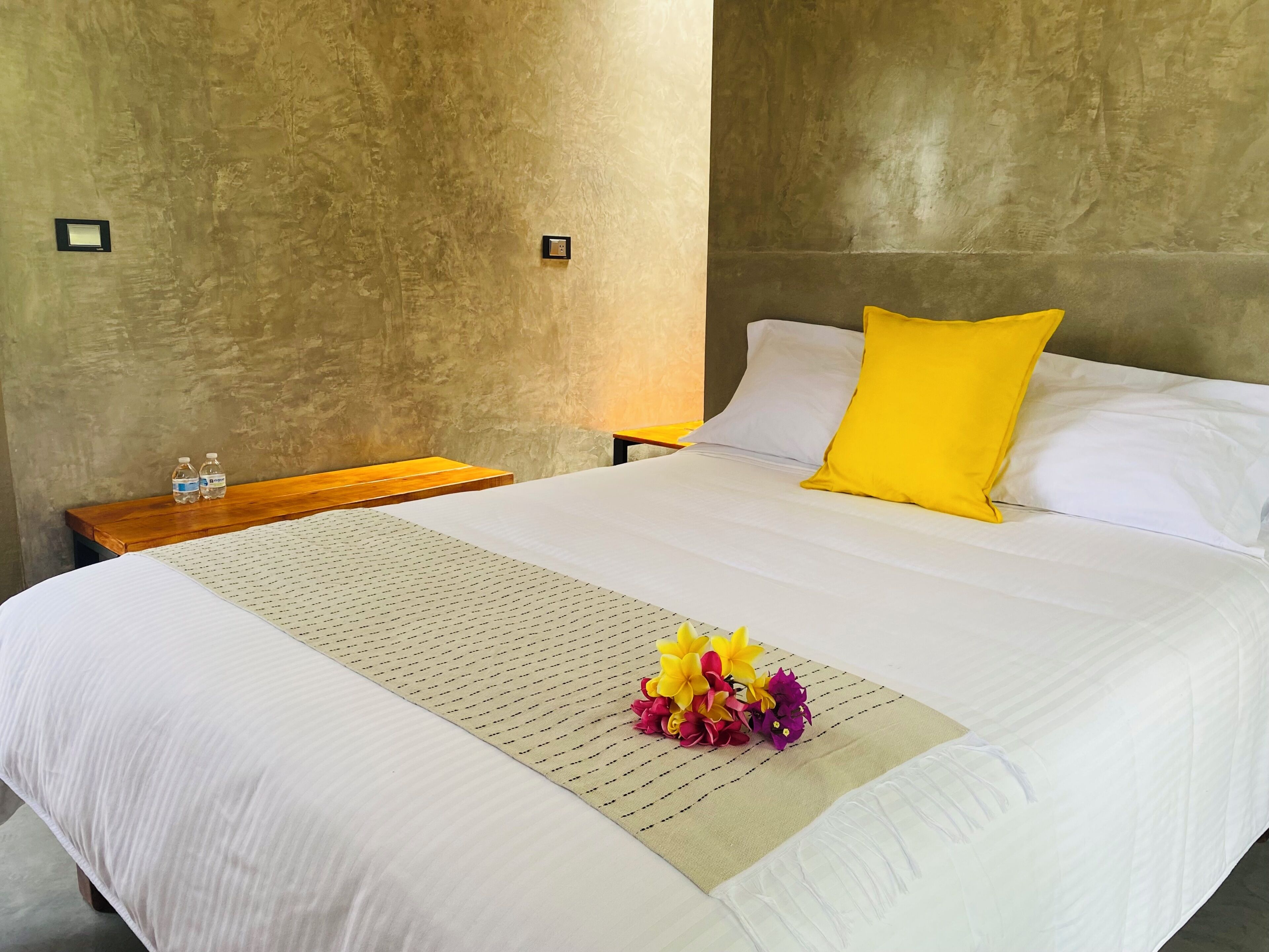 A hotel room with a large bed, a wooden nightstand, a water bottle, and a flower arrangement.