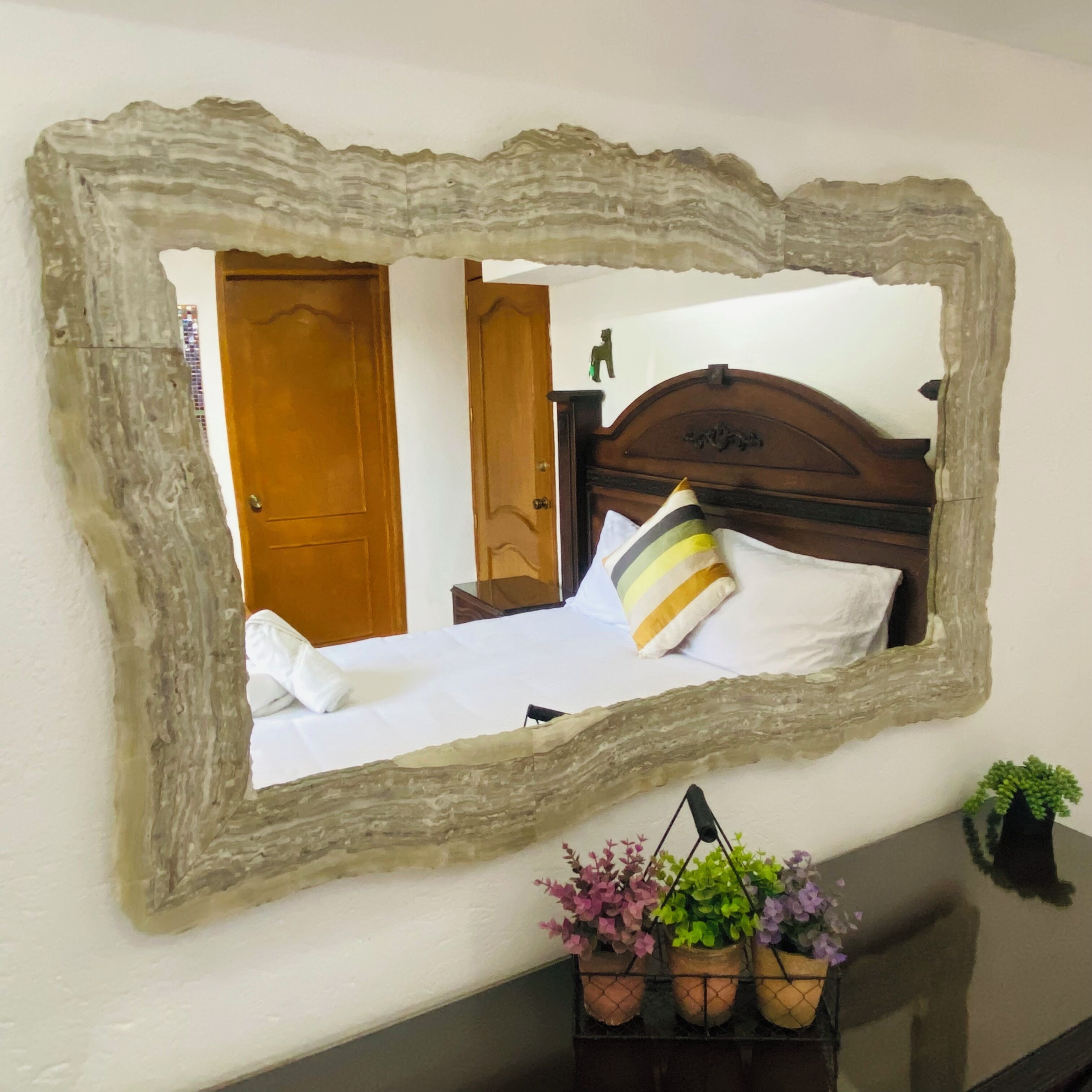 A bedroom with a wooden headboard, a bed with white bedding, a mirror with a rustic frame, and a small plant on a nightstand.