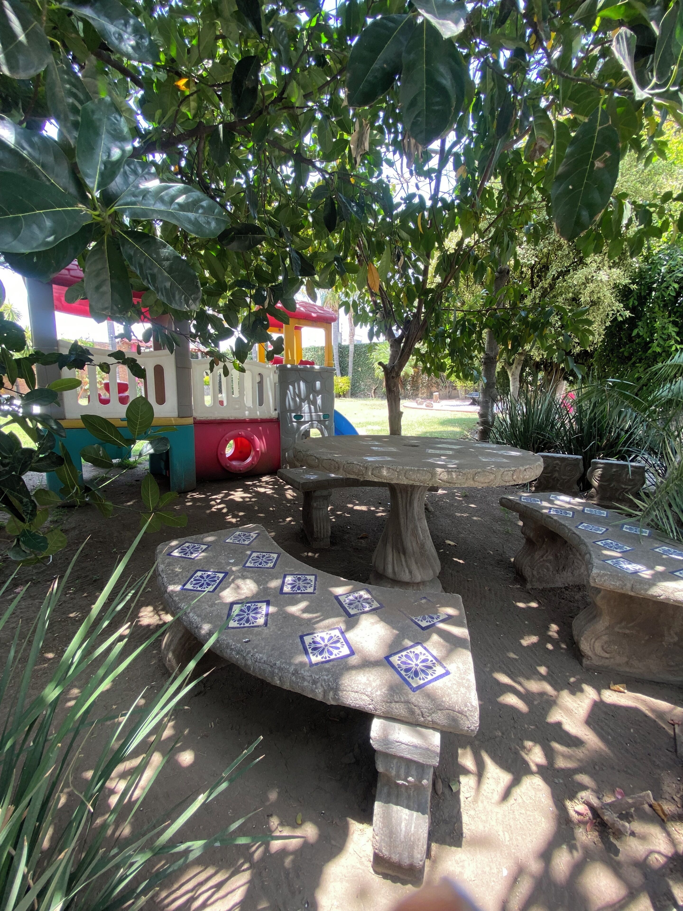 Un parque con área de juegos infantiles, bancos de piedra y un árbol.