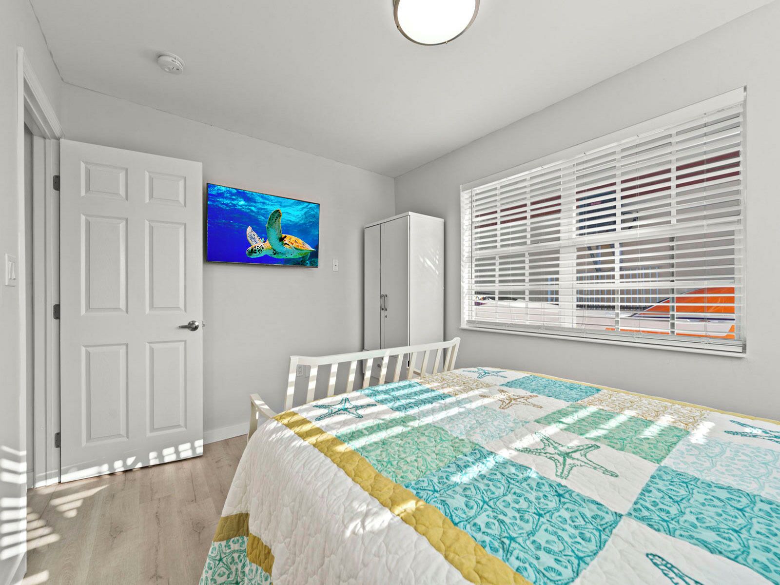 A bedroom with a bed, a TV on the wall, a window with blinds, and a door.