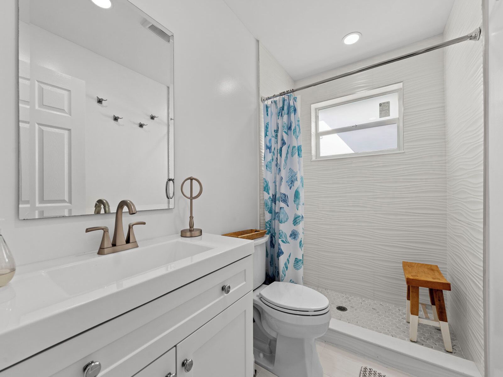 Une salle de bain avec un lavabo blanc, deux robinets, un miroir, des toilettes et une douche équipée d’un tabouret en bois.