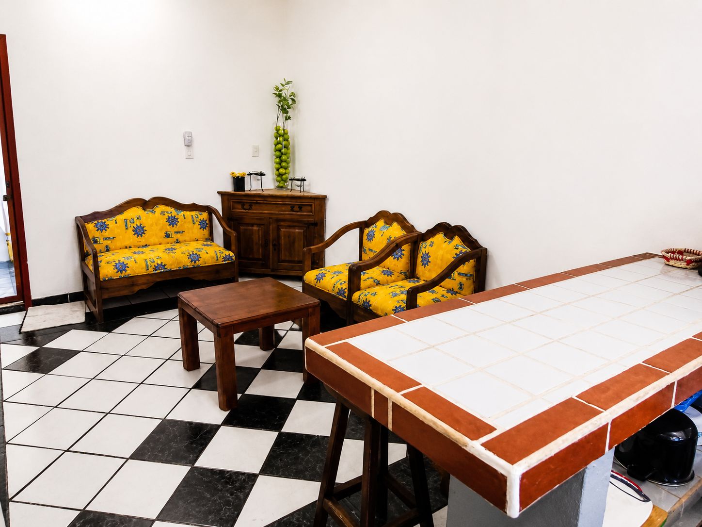Una habitación con piso a cuadros, dos sofás de madera con cojines amarillos, una mesa de madera y una mesita de centro con baldosas blancas y marrones.