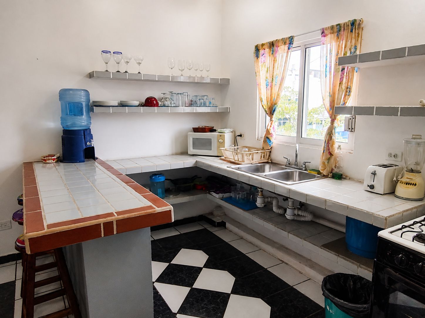 A kitchen with a tiled floor, a water dispenser, a microwave, a sink, a stove, and a window with curtains.