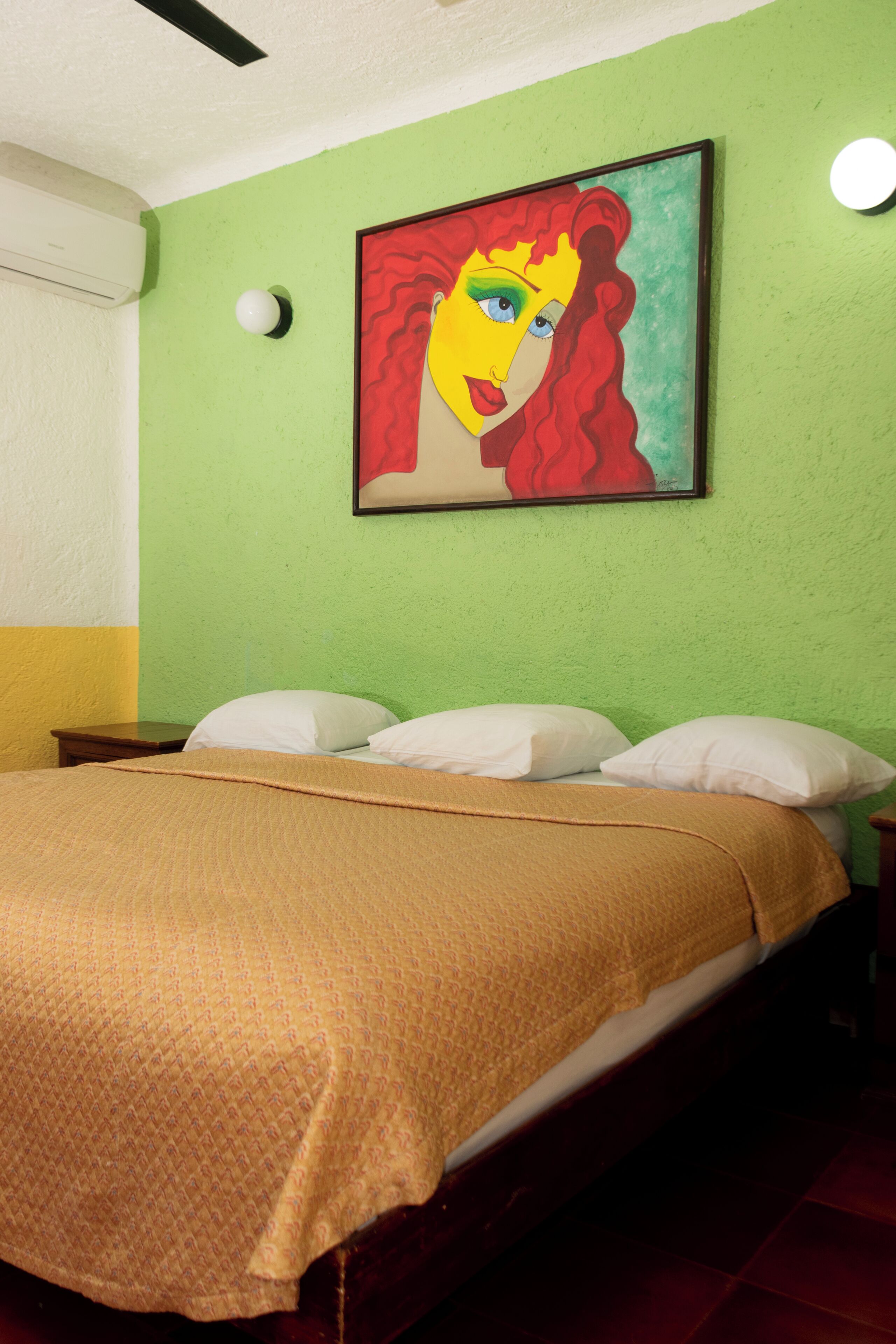 A room with a bed, a green wall, a painting, and a white air conditioning unit.