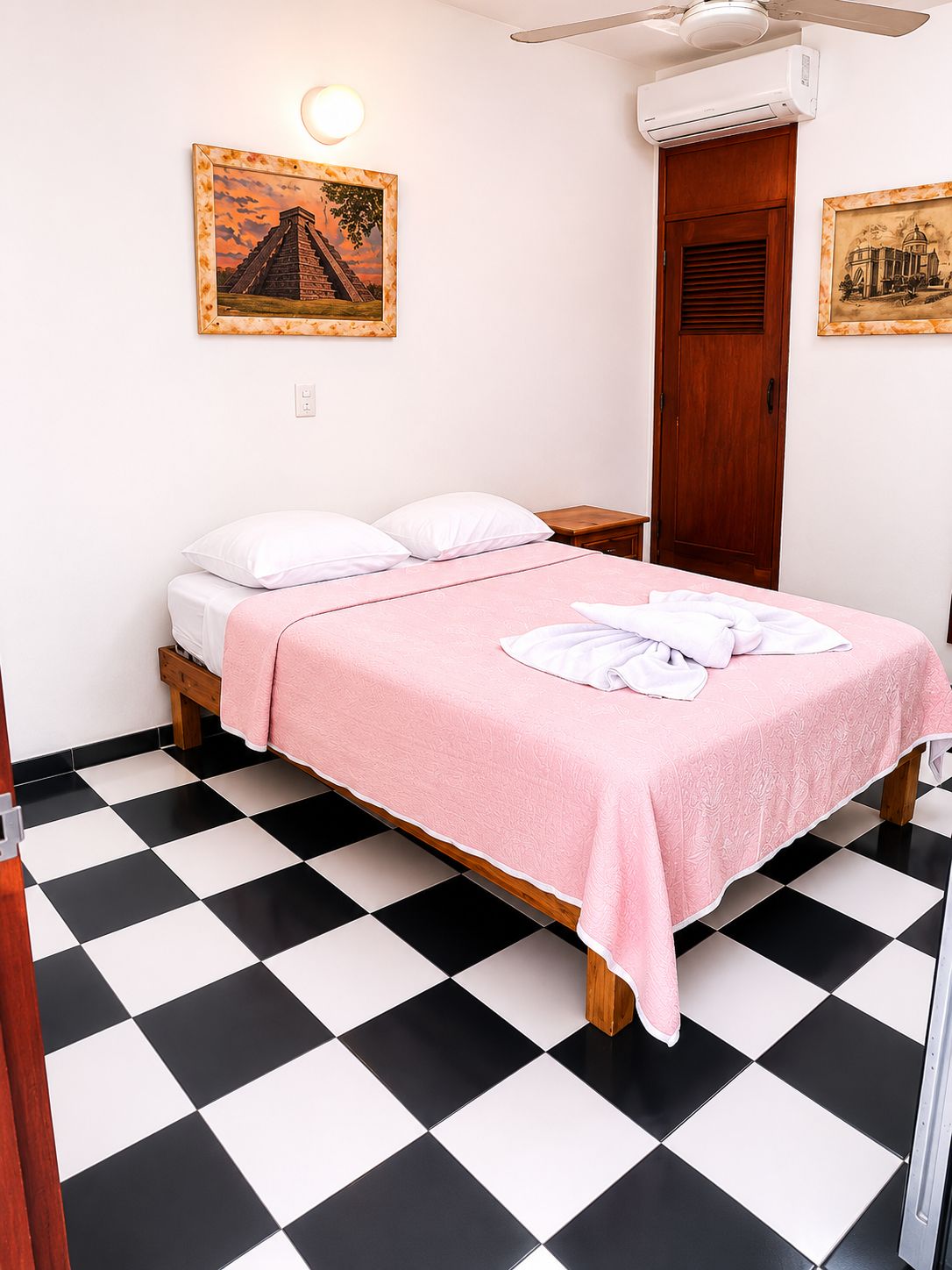 A single bed with a pink bedspread, white pillows, and a white towel on the bed. There are two wall-mounted pictures, one of a pyramid and another of a building. The room has a black and white checkered floor.