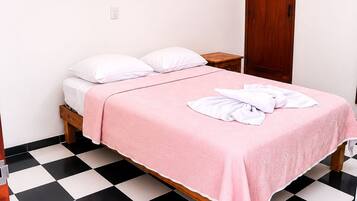 A single bed with a pink bedspread, white pillows, and a white towel on the bed. There are two wall-mounted pictures, one of a pyramid and another of a building. The room has a black and white checkered floor.