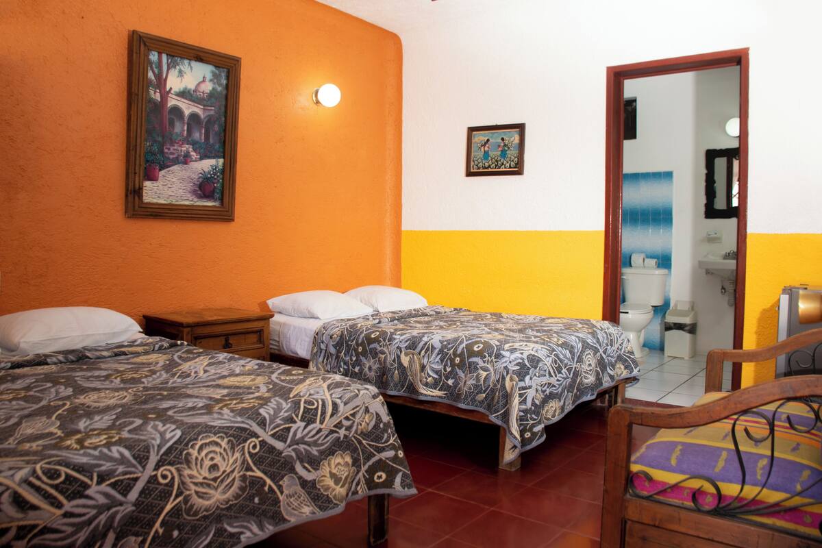 Une chambre avec deux lits, une chaise en bois, un tableau accroché au mur et une salle de bain visible à travers une porte ouverte.