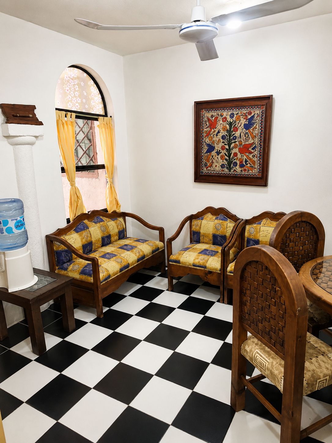Ein Zimmer mit Schachbrettfußboden, zwei Holzsofas mit gemusterten Kissen, einem Deckenventilator, einem Wasserspender und einem gerahmten Kunstwerk an der Wand.