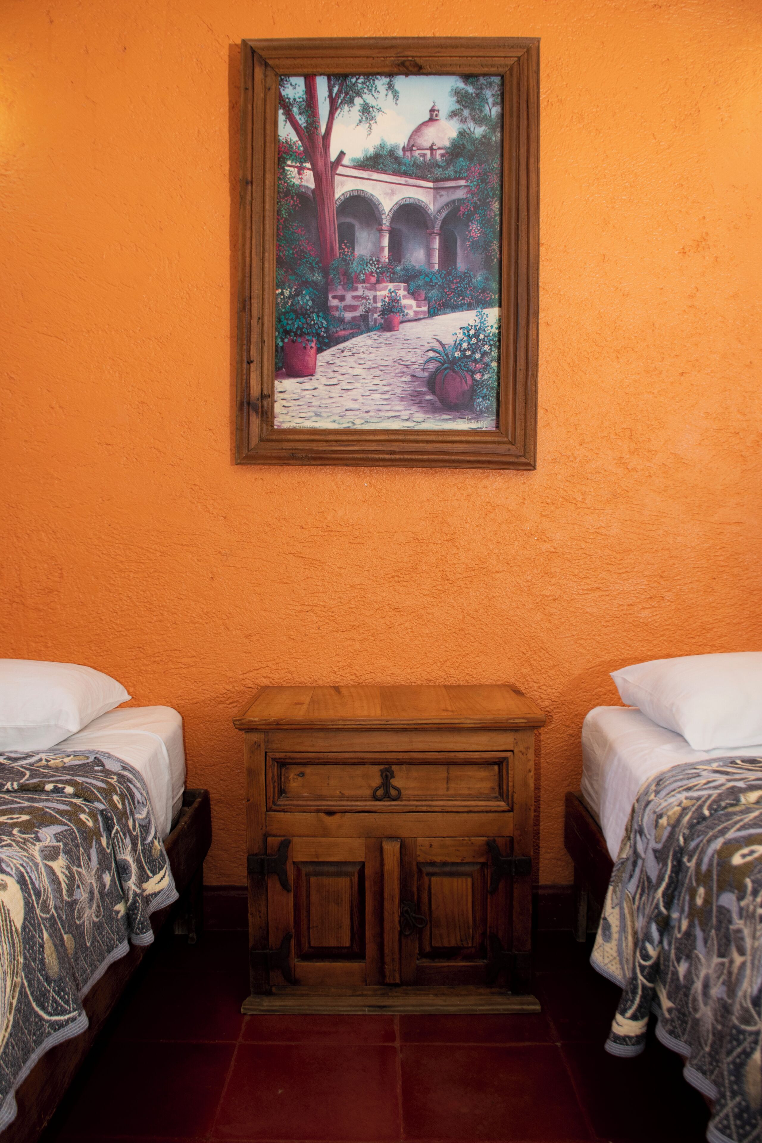 A room with two beds, a wooden nightstand, and a painting of a courtyard.
