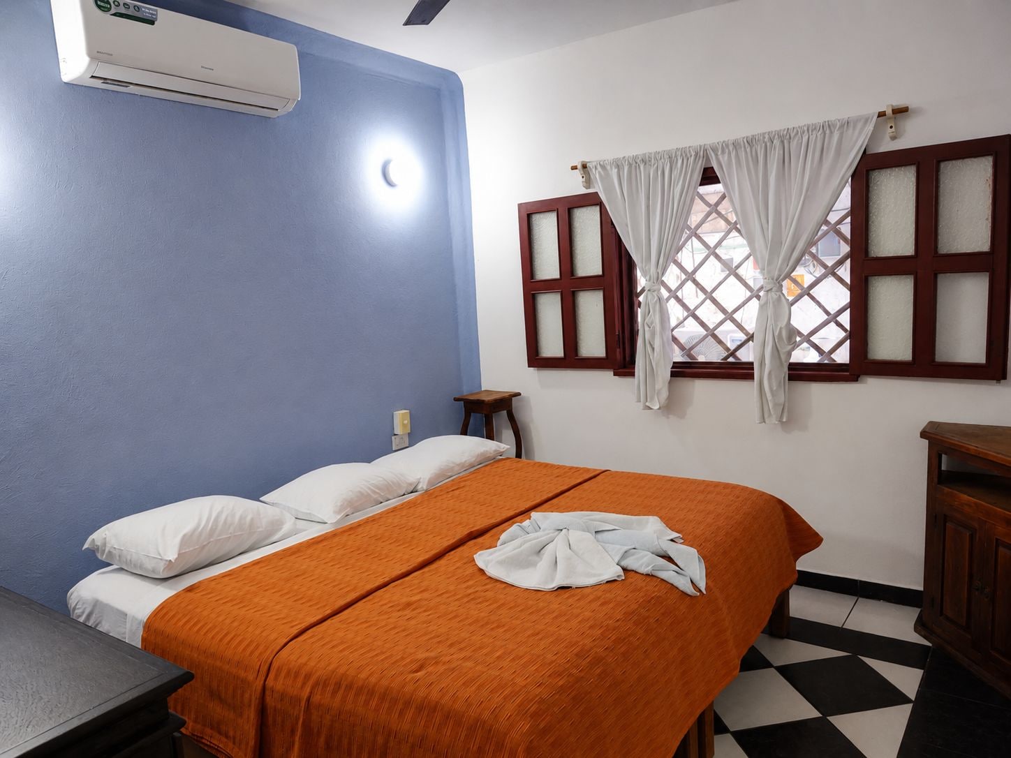 A hotel room with a bed, a nightstand, a window with curtains, and a wall-mounted air conditioner.