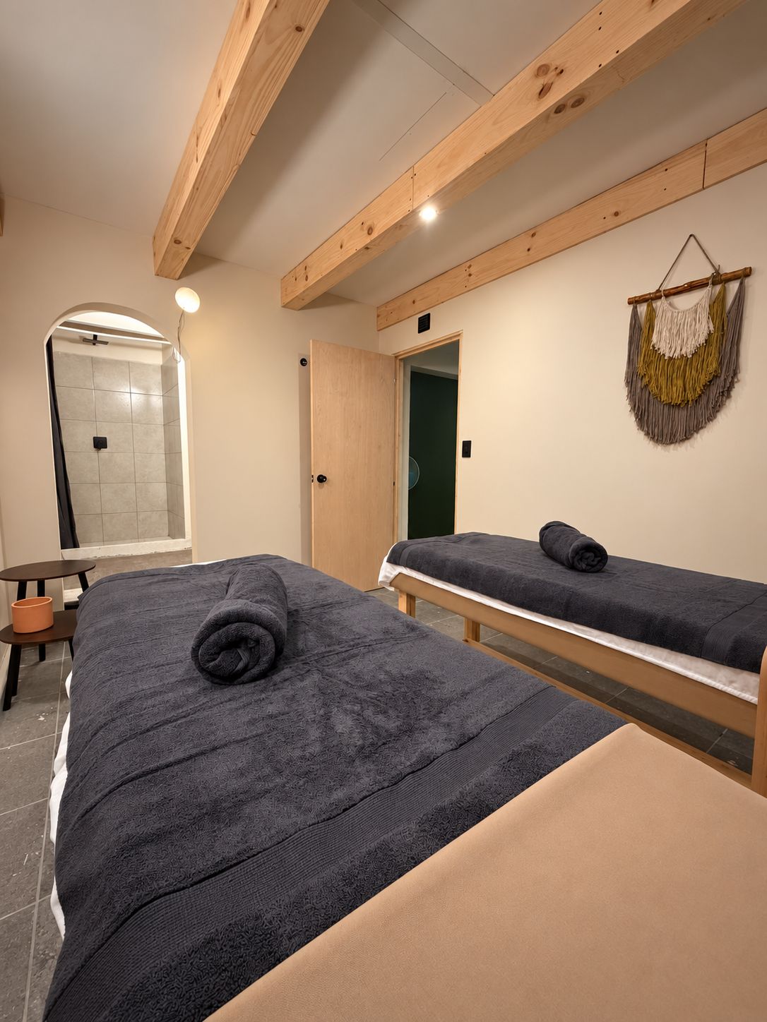 Una habitación con dos camas, una mesita, un banco, un macramé colgado en la pared y una zona de ducha.