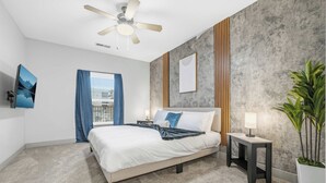 A modern hotel room with a bed, a ceiling fan, a nightstand with a lamp, a large window with curtains, and a plant. - Spacious Modern 2BR 2Bath | Free Parking | Near City Center (Indianapolis)