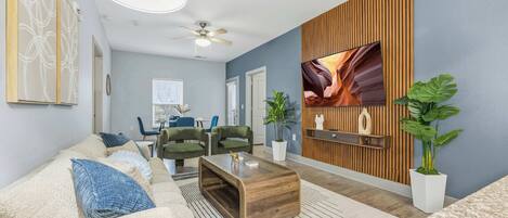 A modern living room with a sofa, coffee table, and chairs, a flat-screen TV mounted on a wooden wall, and a ceiling fan.