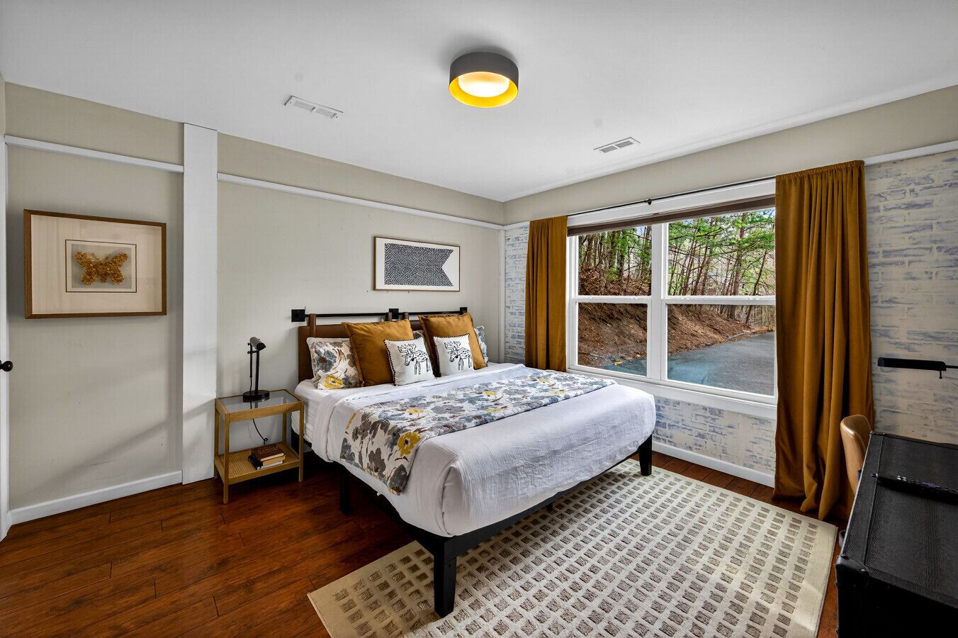 A bedroom with a bed, a nightstand, a lamp, a window with curtains, and a view of the outdoors.