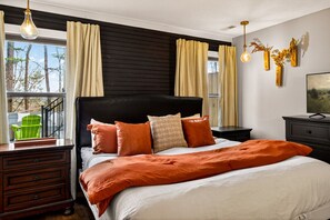 A bedroom with a large bed, dark headboard, and multiple pillows. There is a dresser, a nightstand, and a TV on a dresser. The room has a window with curtains and a view of trees outside. - Dolly`s Outdoor Oasis - Hot Tub, Sauna & More! (Sevierville)