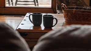 Une pièce chaleureuse avec une table en bois, deux tasses noires, un panier tressé rempli de pommes de pin et une cheminée en pierre.