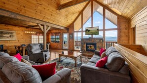 Un intérieur spacieux de chalet en bois, doté d’une cheminée, de sièges confortables et de grandes fenêtres offrant une vue panoramique.
