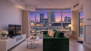 Een moderne woonkamer met een groot raam, een groene bank, een flatscreen-televisie en een kleine tafel met een lamp.
