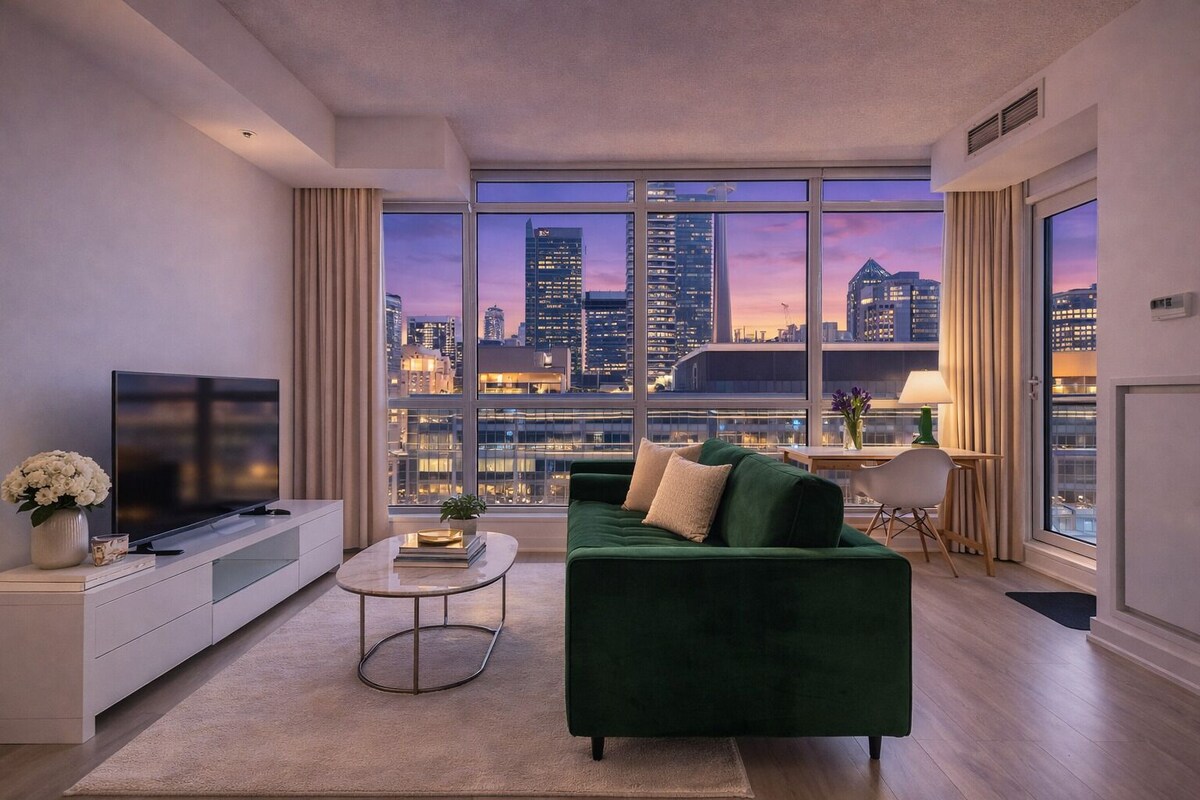 A modern living room with a large window, a green sofa, a flat-screen TV, and a small table with a lamp.