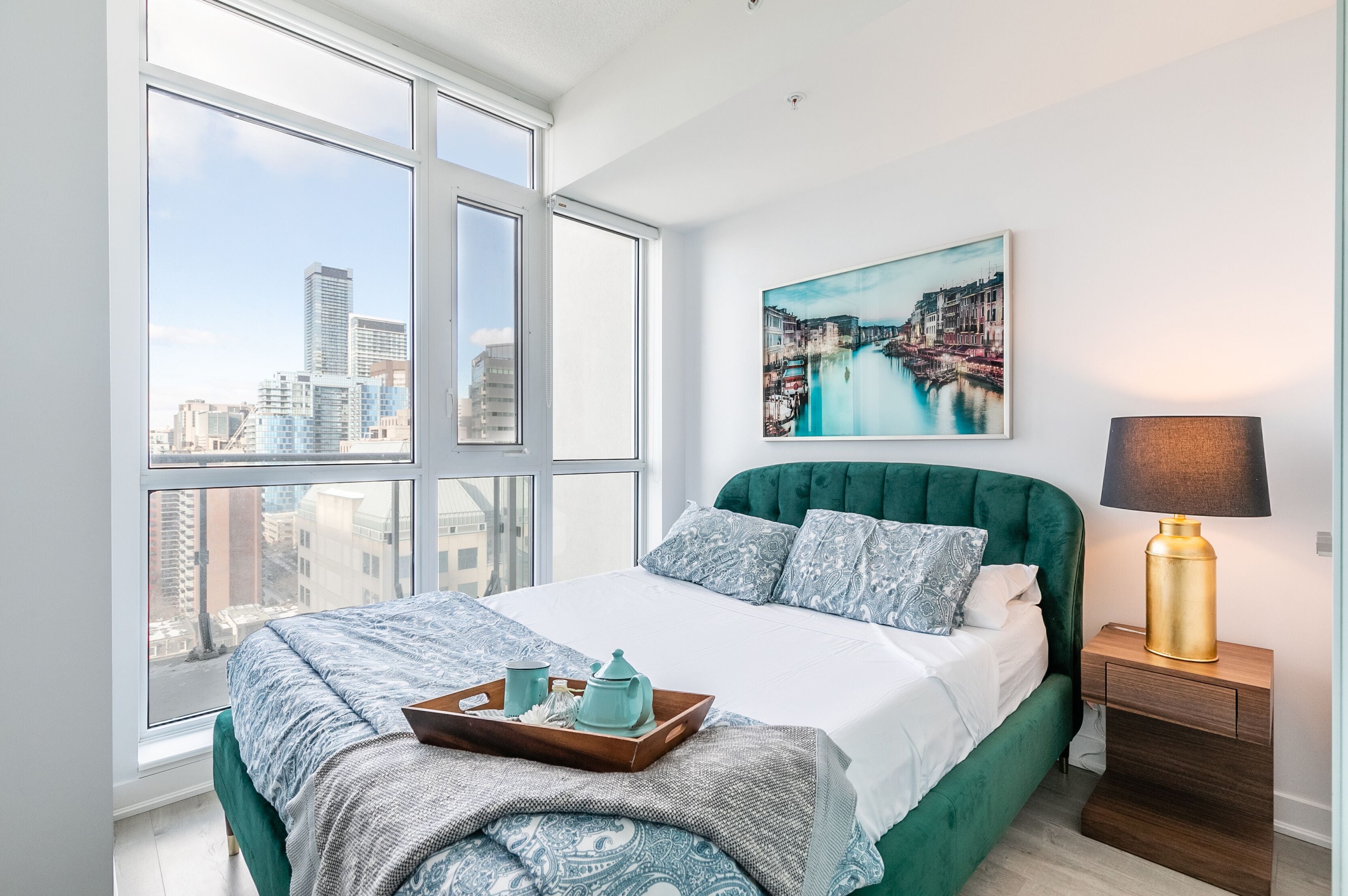 Un dormitorio con un ventanal grande, una cama con ropa de cama verde, vista a la ciudad, un cuadro en la pared y una lámpara de noche.