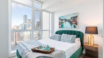 A bedroom with a large window, a bed with green bedding, a city view, a painting on the wall, and a bedside lamp.