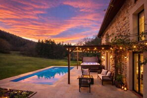 Ein Pool mit Pergola und Sitzgelegenheiten im Freien, ein Steinhaus und ein Sonnenuntergang.