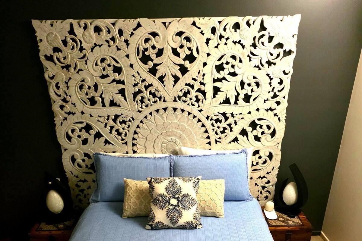 A bed with a blue bedspread and multiple pillows, a decorative headboard, two bedside lamps, and a small wooden chest.