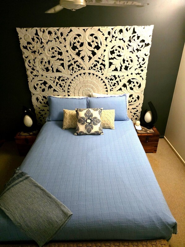 Un lit avec une couvre-lit bleue et plusieurs oreillers, une tête de lit décorative, deux lampes de chevet et une petite commode en bois.