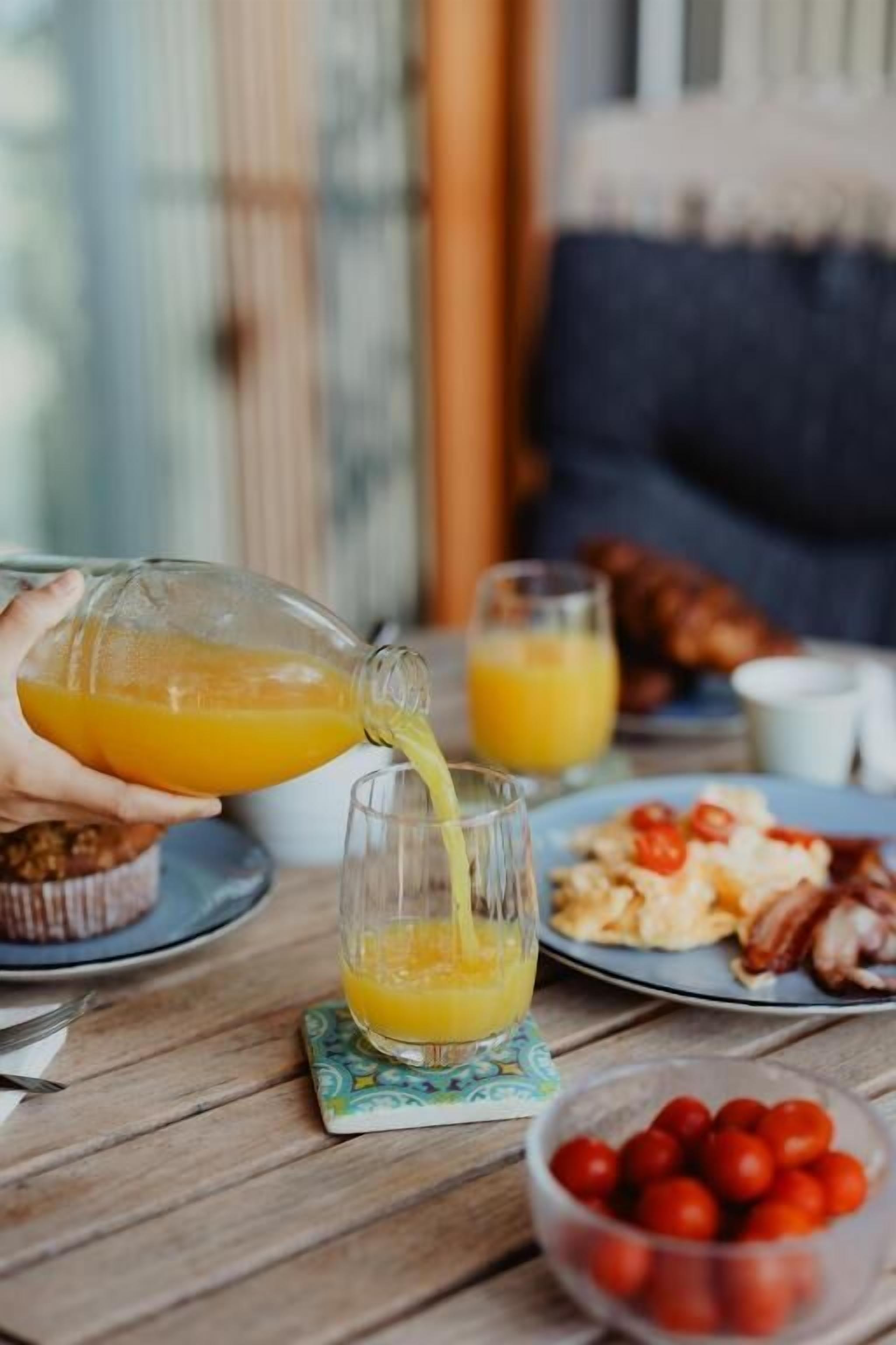 A breakfast setting with a person pouring orange juice into a glass, a plate of eggs and bacon, and a bowl of cherry tomatoes on a wooden table.