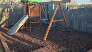 Eine hölzerne Spielkonstruktion mit Rutsche und Schaukeln, umgeben von einem Holzzaun und Rindenmulch.