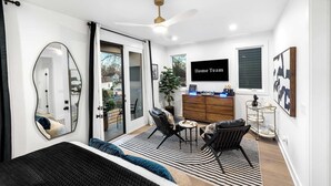 A modern bedroom with a large bed, a ceiling fan, a TV, a dresser, and a dining area with a plant. - Downtown Raleigh Rooftop Retreat - Skyline Views (Raleigh)