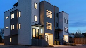 A modern multi-story residential building with exterior lighting, a visible entrance, and a wooden fence in the background. - Downtown Raleigh Rooftop Retreat - Skyline Views (Raleigh)