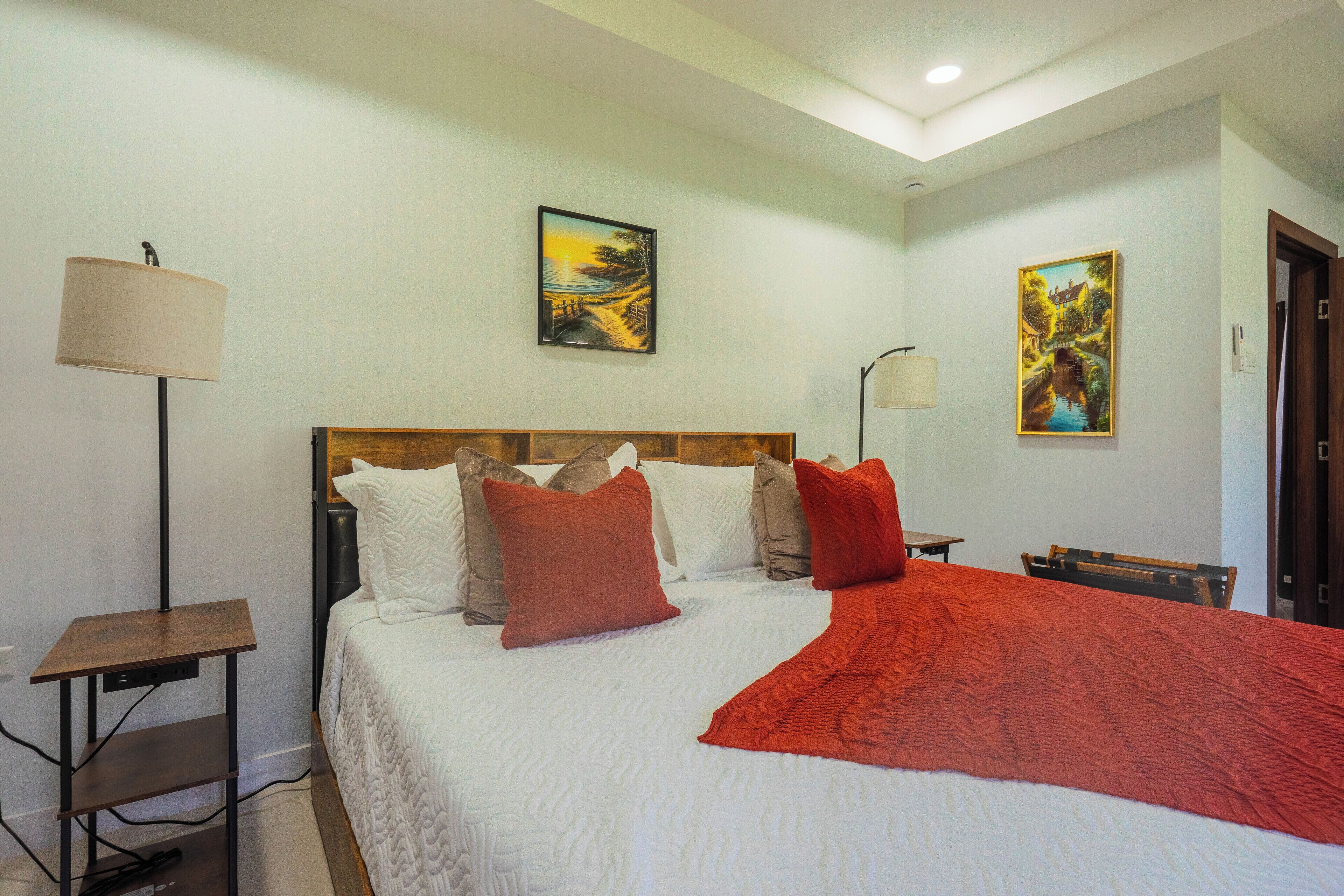 A neatly made bed with a white comforter and red accents, a bedside table with a lamp, and two wall-mounted paintings.