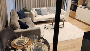 Uma sala de estar moderna com um sofá, uma mesa de jantar posta com pratos, copos e uma vela, e uma área de cozinha com armários e eletrodomésticos.
