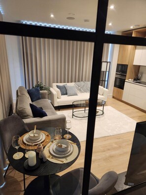 A modern living room with a sofa, a dining table set with plates, glasses, and a candle, and a kitchen area with cabinets and appliances. - Executive Apartment | GX Golf Club | Parking (Chalfont Saint Peter)