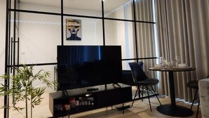 Uma sala de estar moderna com uma televisão de tela plana, uma mesa de jantar com cadeiras, um vaso de planta e uma grande janela com cortinas.